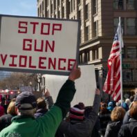 Image shows a small view of a larger protest in a US city. Prominent in the photo are an older white man holding a sign that reads STOP GUN VIOLENCE in red letters against a white background, and the US flag.