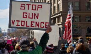 Image shows a small view of a larger protest in a US city. Prominent in the photo are an older white man holding a sign that reads STOP GUN VIOLENCE in red letters against a white background, and the US flag.