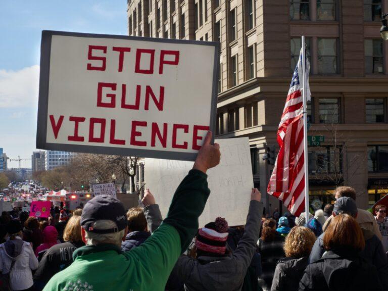 Image shows a small view of a larger protest in a US city. Prominent in the photo are an older white man holding a sign that reads STOP GUN VIOLENCE in red letters against a white background, and the US flag.