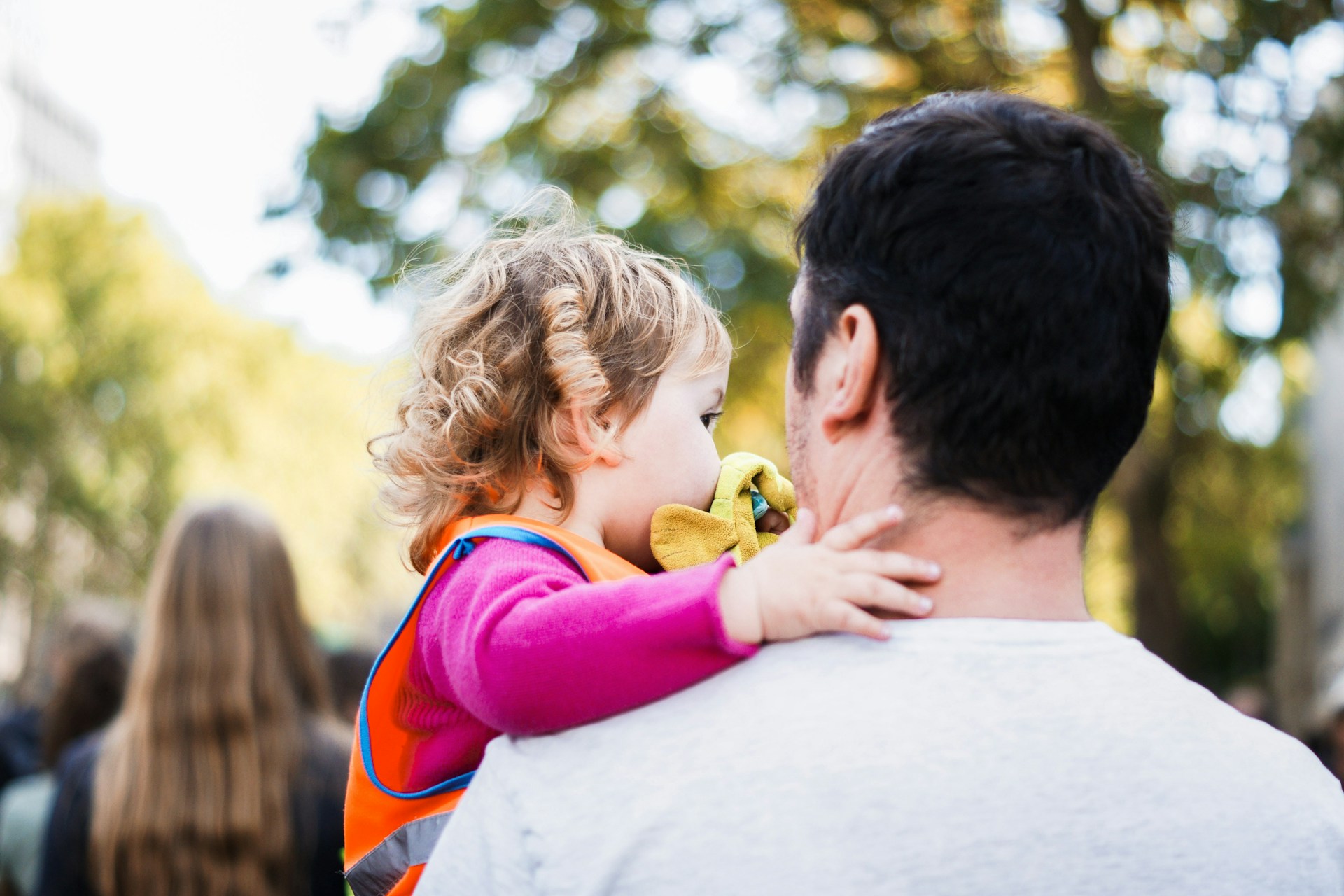 image shows a toddler with blonde curly hair wearing a fuchsia sweater being held by a man with short dak hair wearing a white t-shirt. The perspective is from behind the man at his left, so we see the right side of the child's face. In the background is a natural setting with a large tree on a warm, sunny day.