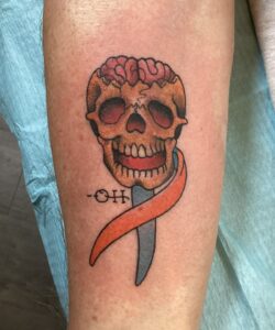 Image shows a human forearm with a fresh tattoo of a human skull with out a cranium, exposing the brain, and with a full set of teeth that make the skull appear to be smiling. Wrapped around what would be the neck of the skull are two ribbons: one orange and one green. Just left of center near the chin of the skull, we see the letters - O H.