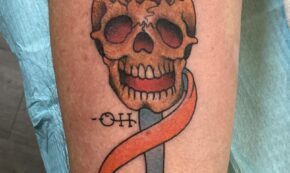 Image shows a human forearm with a fresh tattoo of a human skull with out a cranium, exposing the brain, and with a full set of teeth that make the skull appear to be smiling. Wrapped around what would be the neck of the skull are two ribbons: one orange and one green. Just left of center near the chin of the skull, we see the letters - O H.