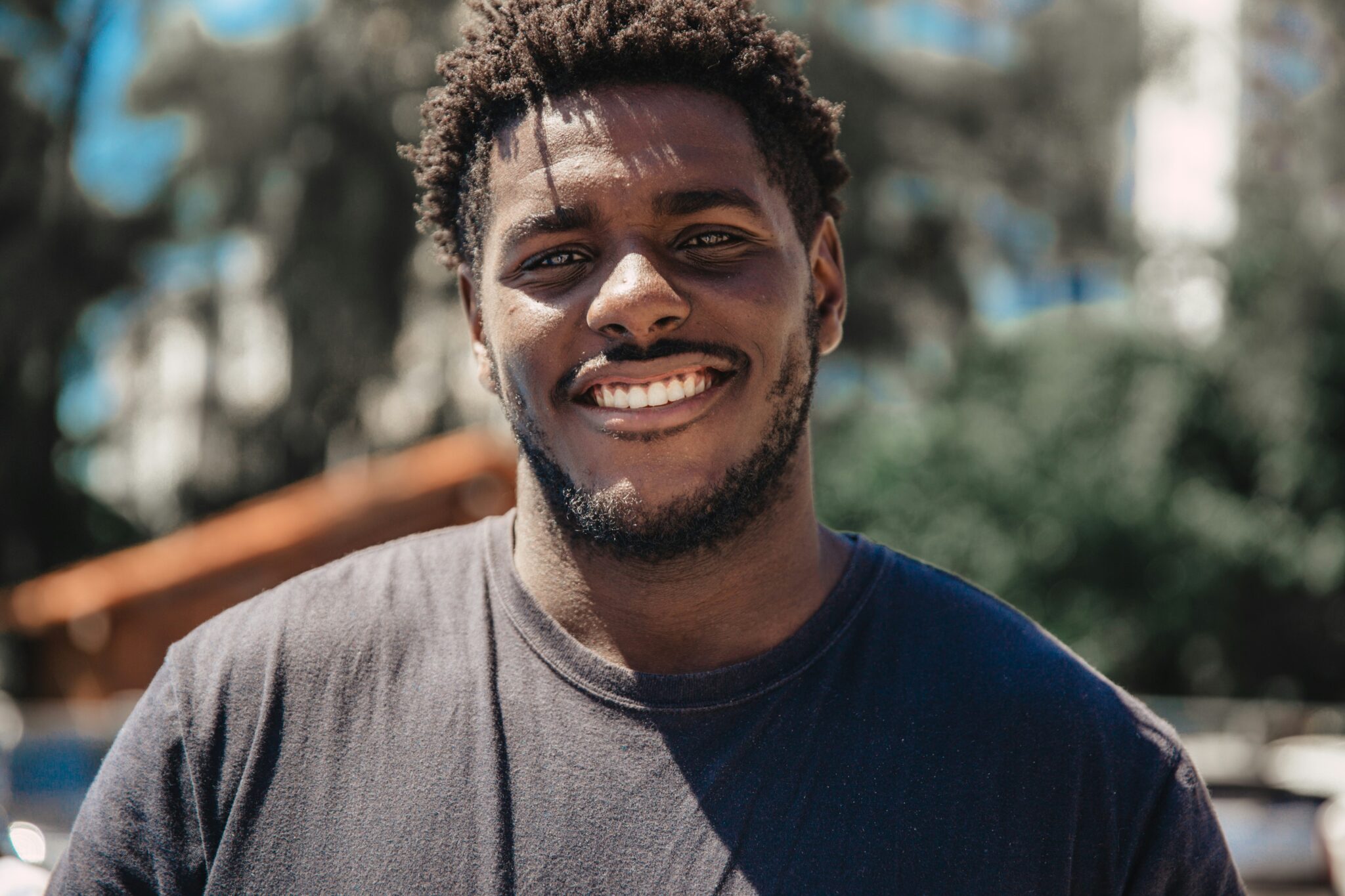30-ish y.o. Black man with short twists and a big white smile wearing a black t-shirt outside on a sunny day.