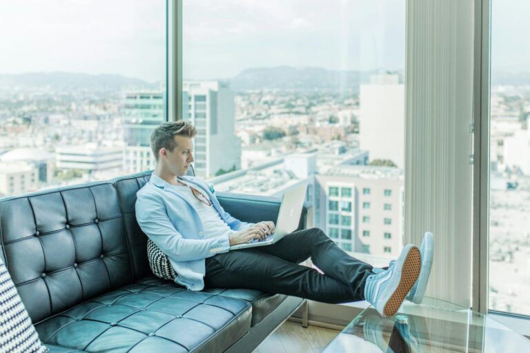 Image shows a young male with light skin and blond hair sitting on a tufted black leather couch in a high-rise corner office suite. He is wearing a light blue blazer and matching high-top sneakers with straight-leg jeans. He is reading on an Apple laptop and has his feet propped up on the coffee table.