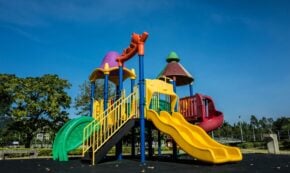 Photo shows a colorful playground against a deep blue sky with large deciduous trees in the background.
