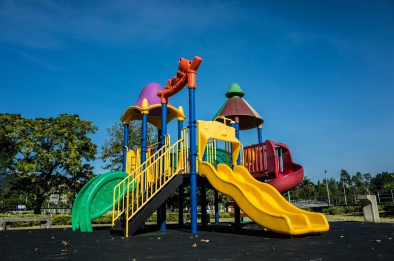 Photo shows a colorful playground against a deep blue sky with large deciduous trees in the background.