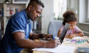 Father working on a laptop while his daughter draws beside him at home